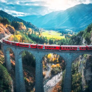 Aerial view of modern red train on Landwasser viaduct in alpine mountains, orange forest at sunset in autumn. Bernina Express, Switzerland. Top view of train in Alps, railroad, colorful trees in fall