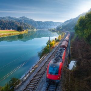 Aerial view of red modern high speed train moving near river in alpine mountains in fog at sunrise in autumn. Top view of train, railroad, lake, reflection, trees in fall. Railway station in Slovenia