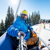 Skier wearing skis, helmet and mask sitting in ski lift cabin taking a selfie with a camera on monopod. Blue sky, sun, winter forest, ski slope with skiers on the blurred background lifestyle concept