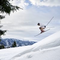 Side view of male skier making jump while sliding down snow-covered slope on skis under beautiful cloudy sky. Man freerider in helmet skiing on fresh powder snow in mountains.