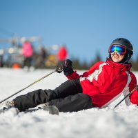 Woman wearing ski goggles, helmet, red jacket, gloves and pants lying with skis on snowy at mountain top and looking away in sunny day with ski lifts and blue sky in background. Carpathian mountains